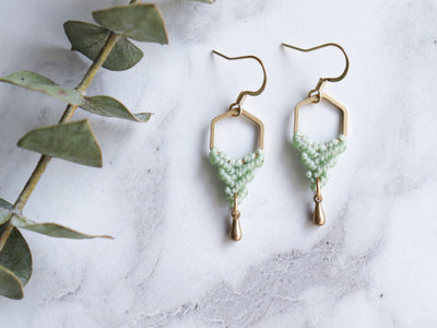 Hexagon shaped drop macrame earrings in green and golden color with white background. 